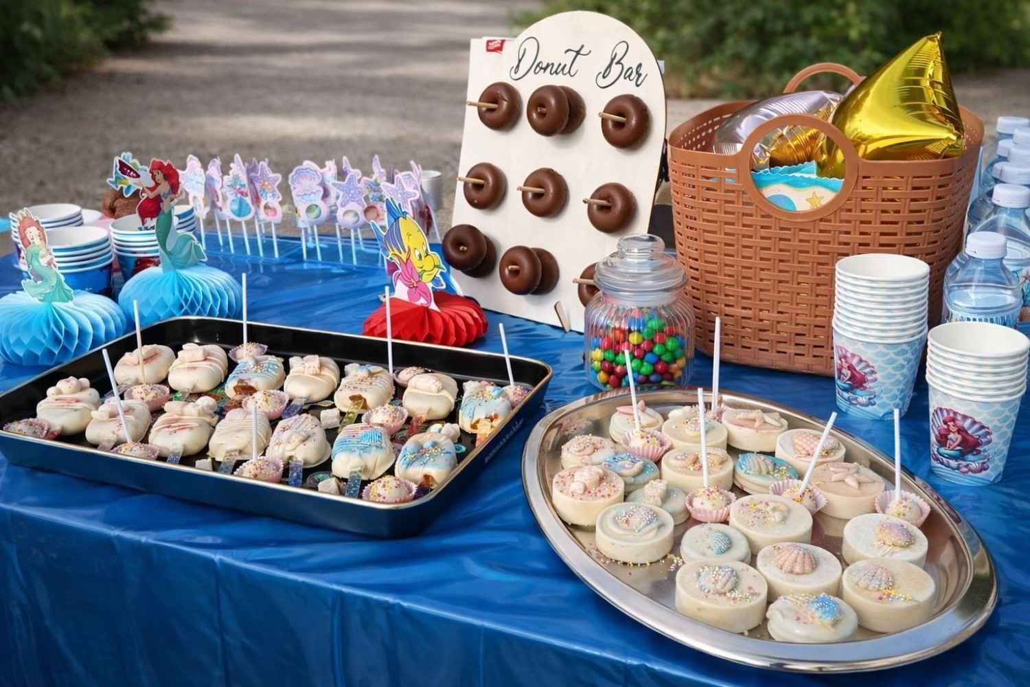 Una mesa azul preparada para una fiesta con un soporte para donas, bandejas de dulces, vasos de papel, una cesta y decoración con temática de sirenas.