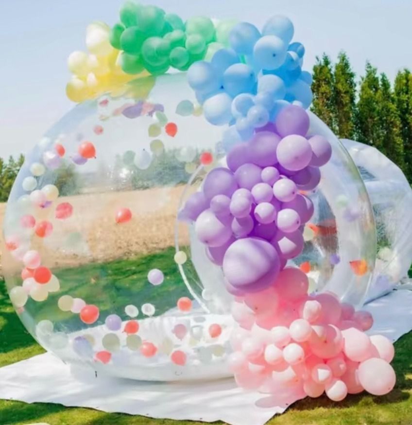 Una carpa inflable transparente con forma de cúpula, decorada con un vibrante arcoíris de globos, instalada sobre un césped.