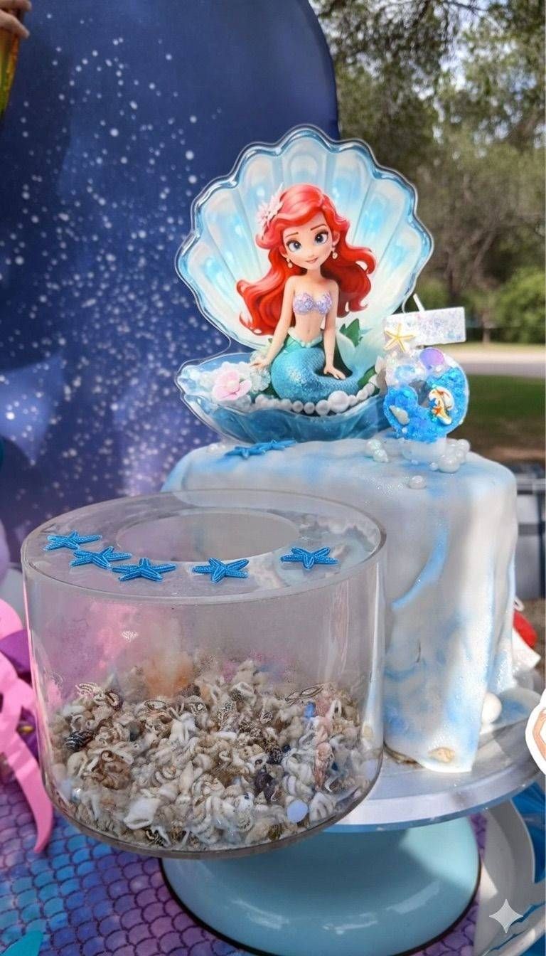 Una tarta de dos pisos con una figurita de Ariel en la parte superior, dentro de una concha marina, y una base transparente rellena de conchas marinas y estrellas azules.