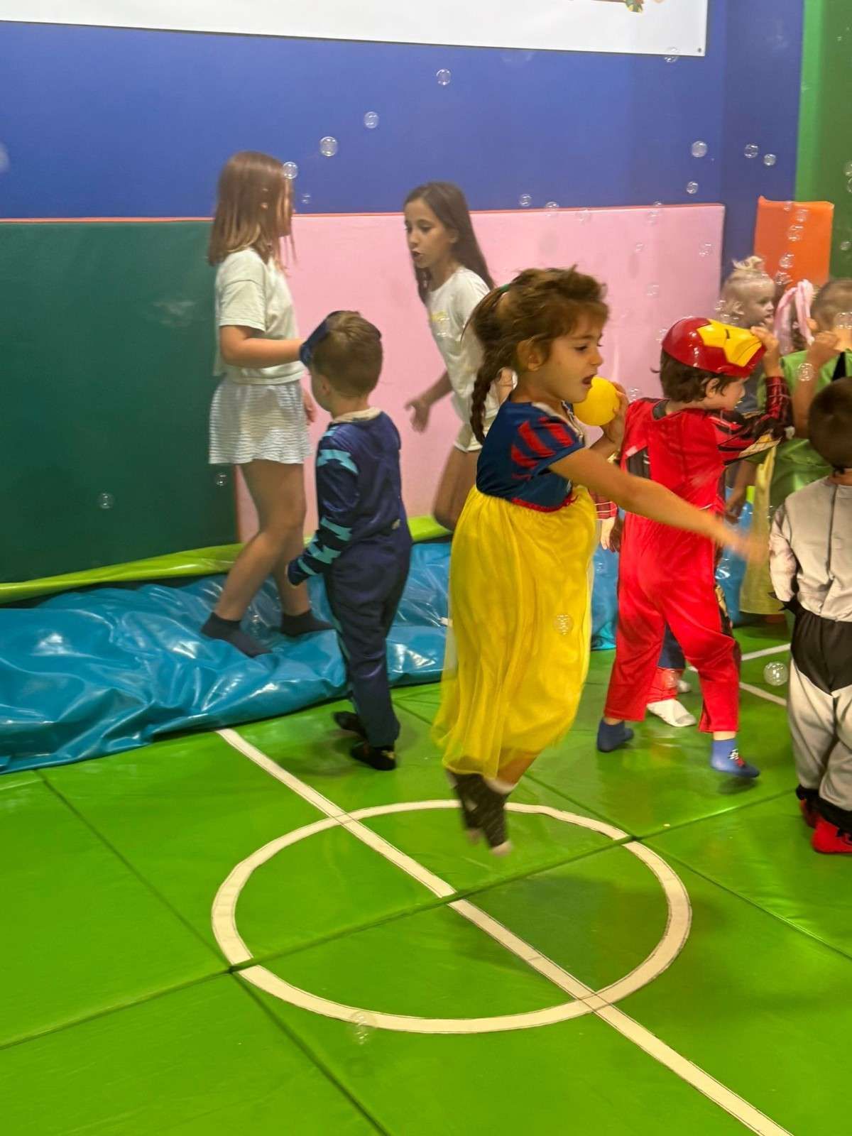 Niños disfrazados juegan en una zona de recreo interior con suelo acolchado verde y paredes azules.