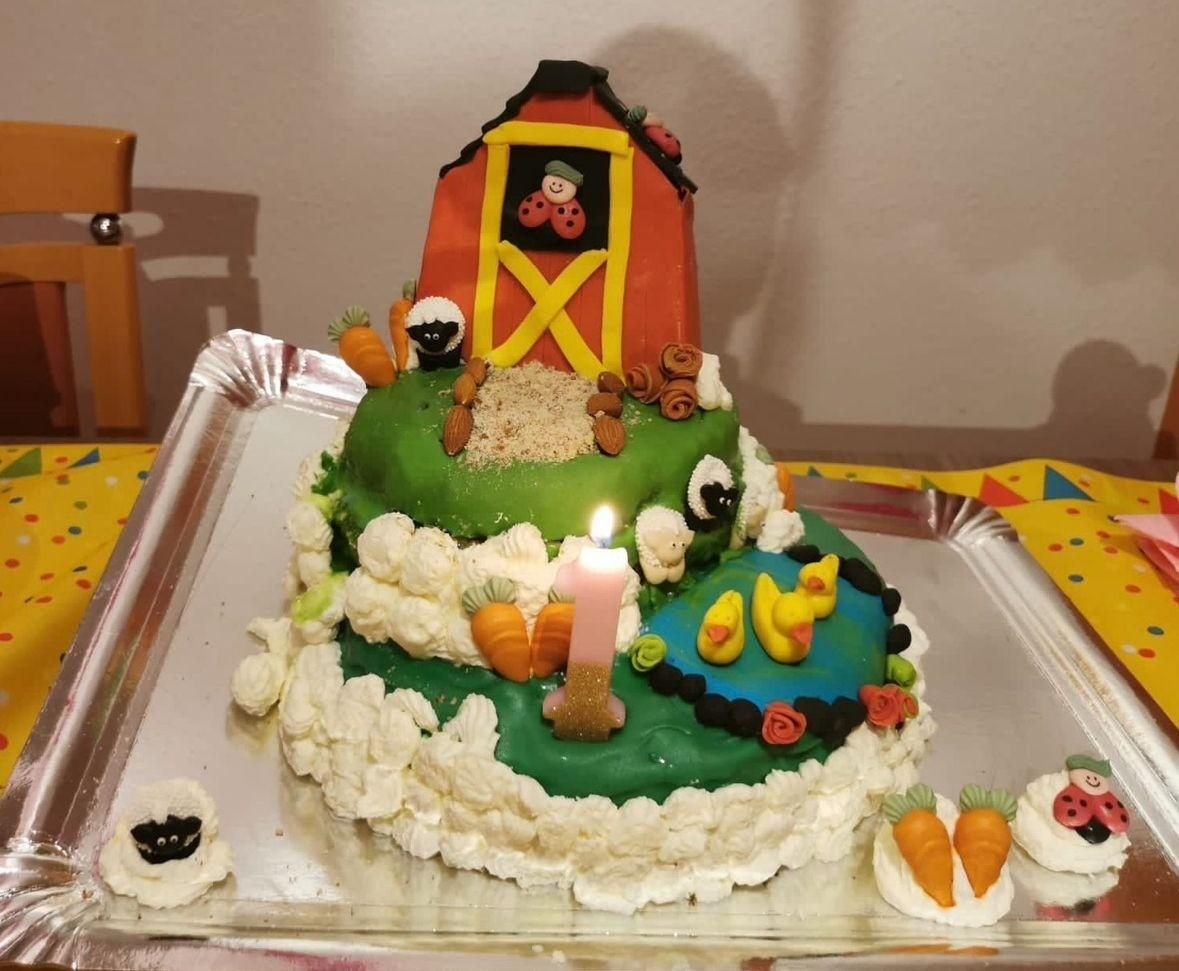 Una tarta de cumpleaños de dos pisos con temática de granja, con un granero, animales de fondant, un estanque con patos y una vela con forma del número 1.