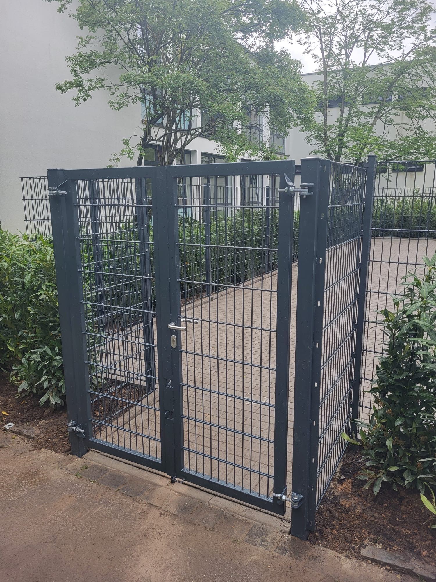 Ein schwarzer Zaun mit einem Tor ist von Bäumen und Büschen umgeben.