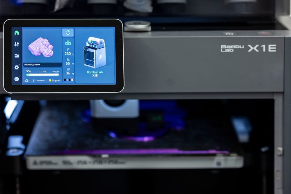 Ein 3D-Drucker mit Touchscreen-Display zeigt ein Modell an und druckt ein violett beleuchtetes Objekt. Die Maschine trägt die Bezeichnung „Ember Lab X1E“.