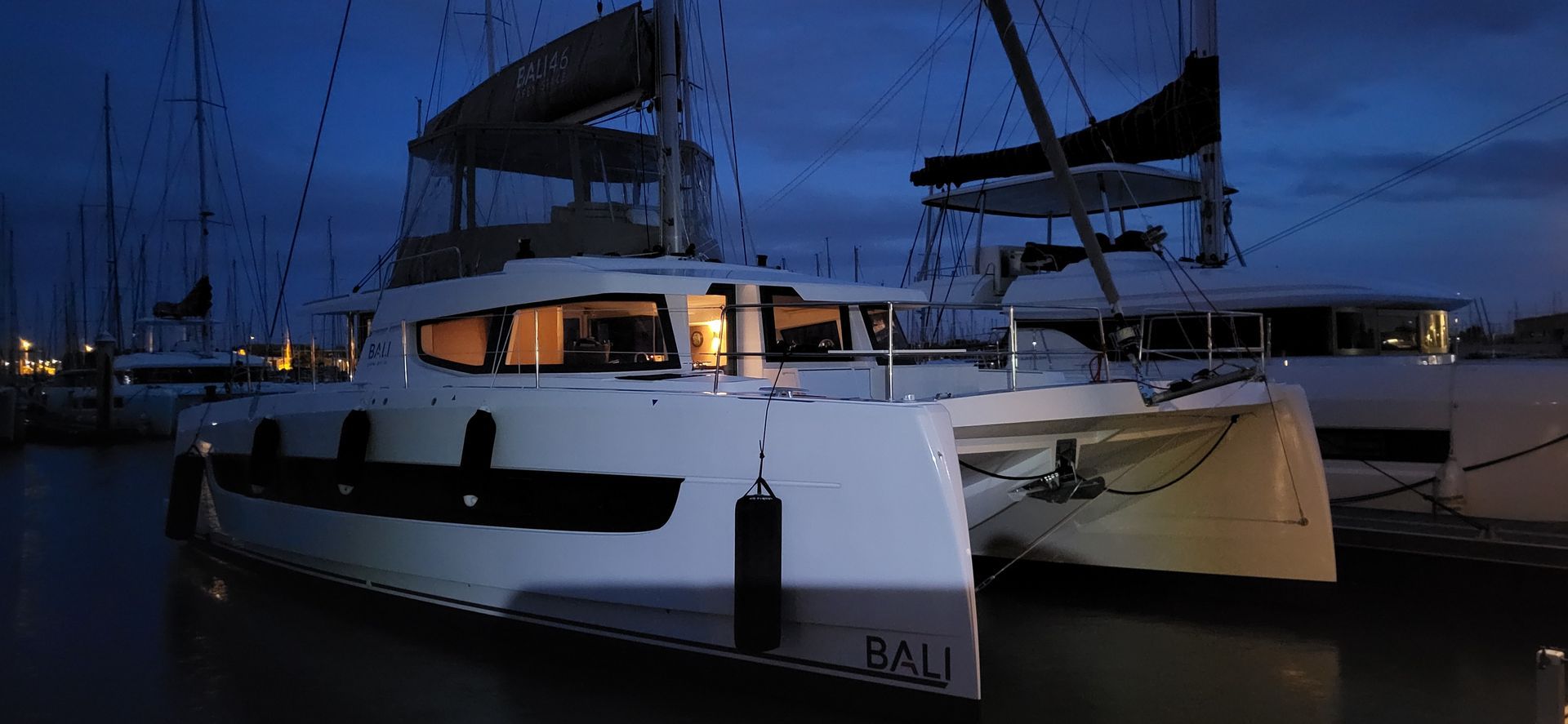 Un catamaran blanc amarré de nuit, fenêtres éclairées, mâts visibles.