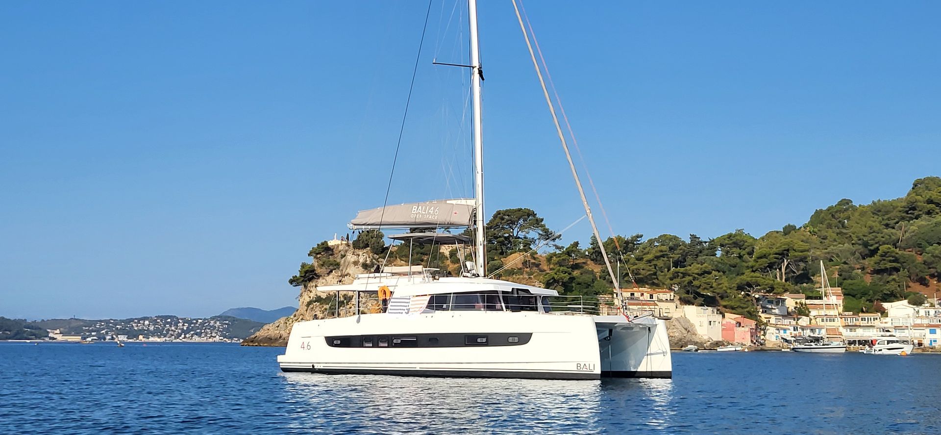 Un catamaran blanc navigue sur une eau bleue près d'une ville côtière entourée d'arbres verdoyants et sous un ciel dégagé.