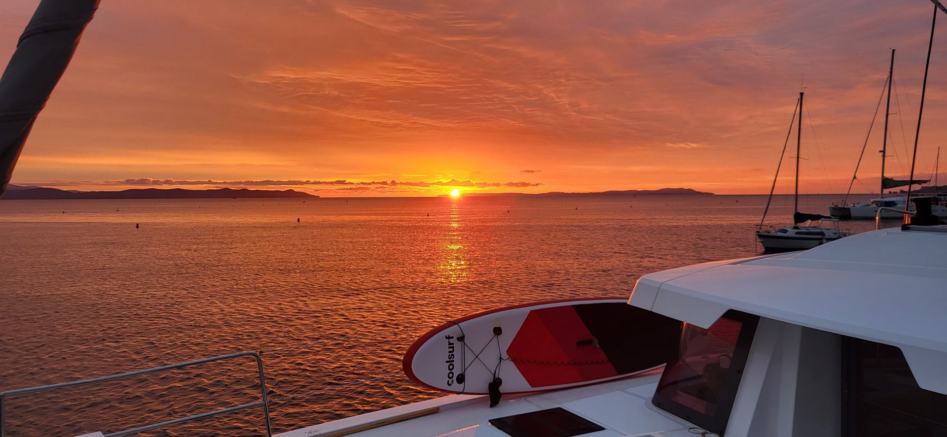 Coucher de soleil sur une mer calme, vu depuis le pont d'un voilier avec une planche à pagaie ; d'autres voiliers au loin.