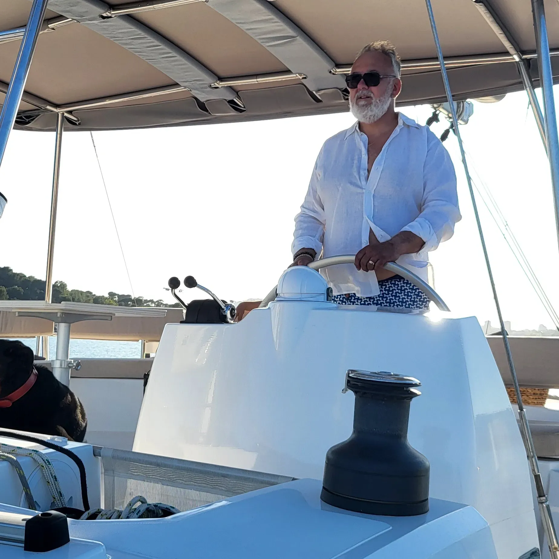 Un homme à la barre d'un voilier blanc, portant des lunettes de soleil et une chemise blanche.