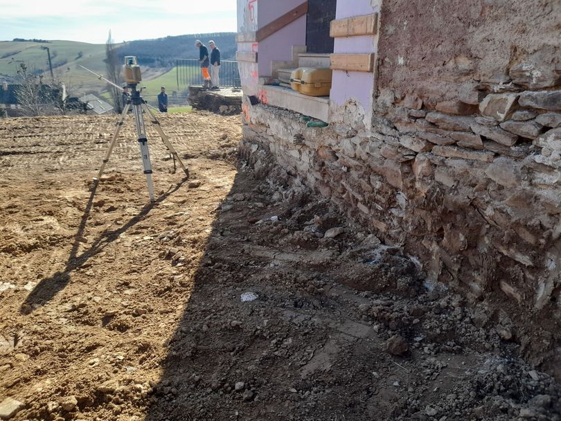 Chantier de construction en milieu rural, jouxtant un mur de pierre, avec des ouvriers et des engins de terrassement en arrière-plan.