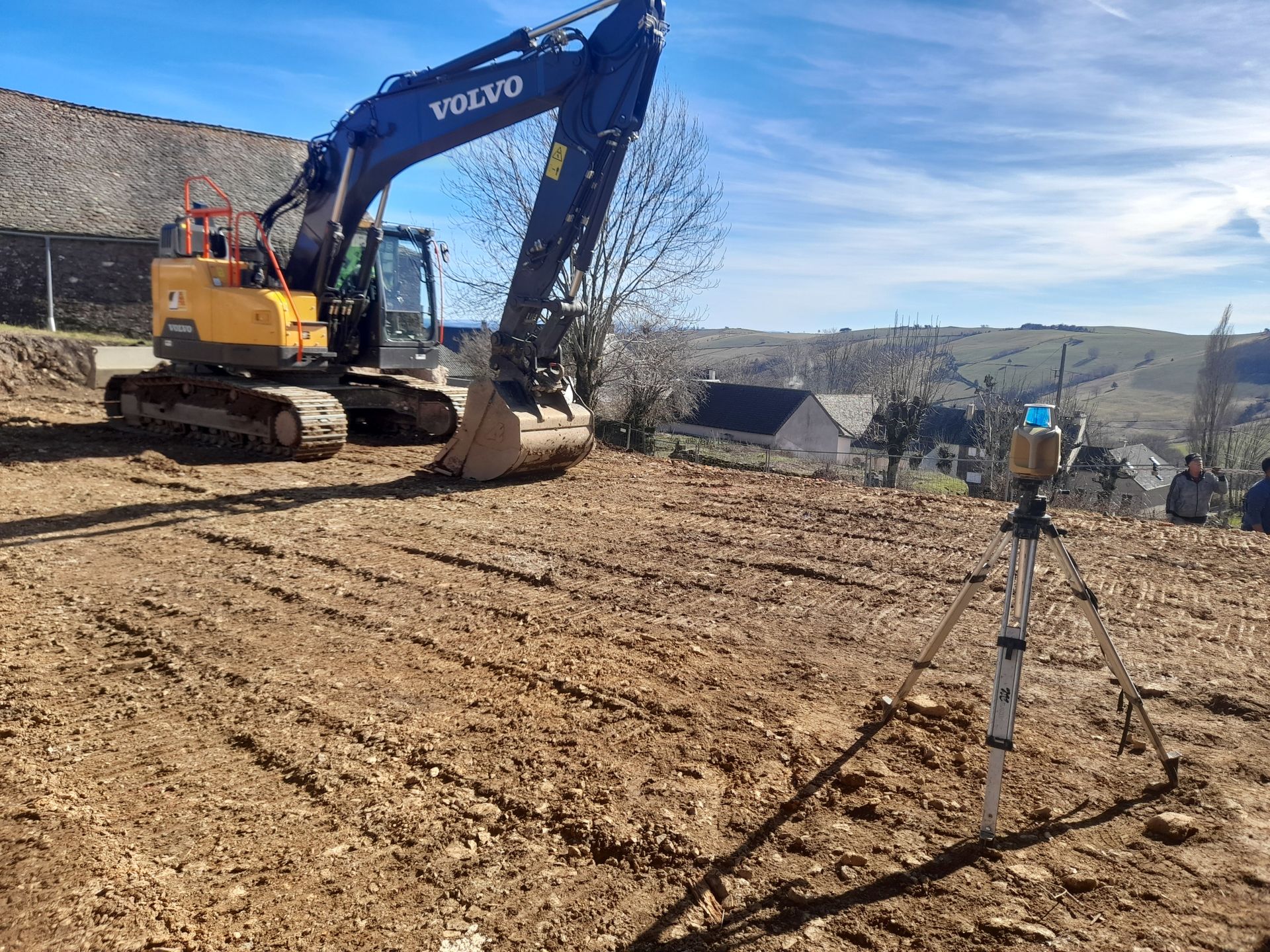Une pelle mécanique Volvo bleue nivelle un terrain vague avec une borne topographique au premier plan.