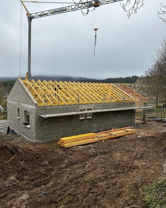 Un chantier de construction où l'on aperçoit un bâtiment en maçonnerie dont la charpente jaune est installée par une grue sous un ciel nuageux.