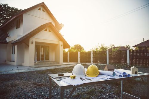 Des casques de chantier blancs et jaunes sont posés sur une table, à côté de plans et d'une maquette de maison, devant une maison de banlieue au coucher du soleil.
