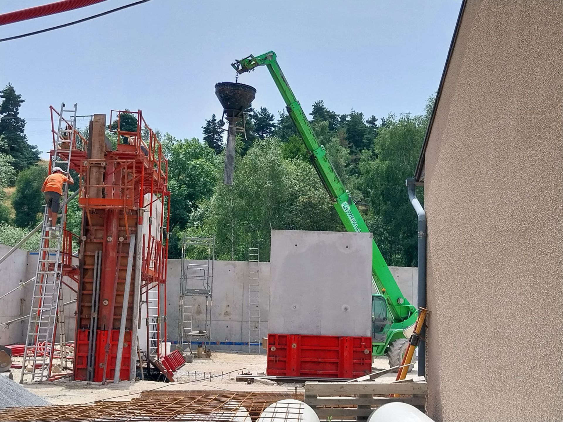 Chantier de construction avec une grue verte soulevant un godet à côté d'échafaudages rouges et de barrières en béton