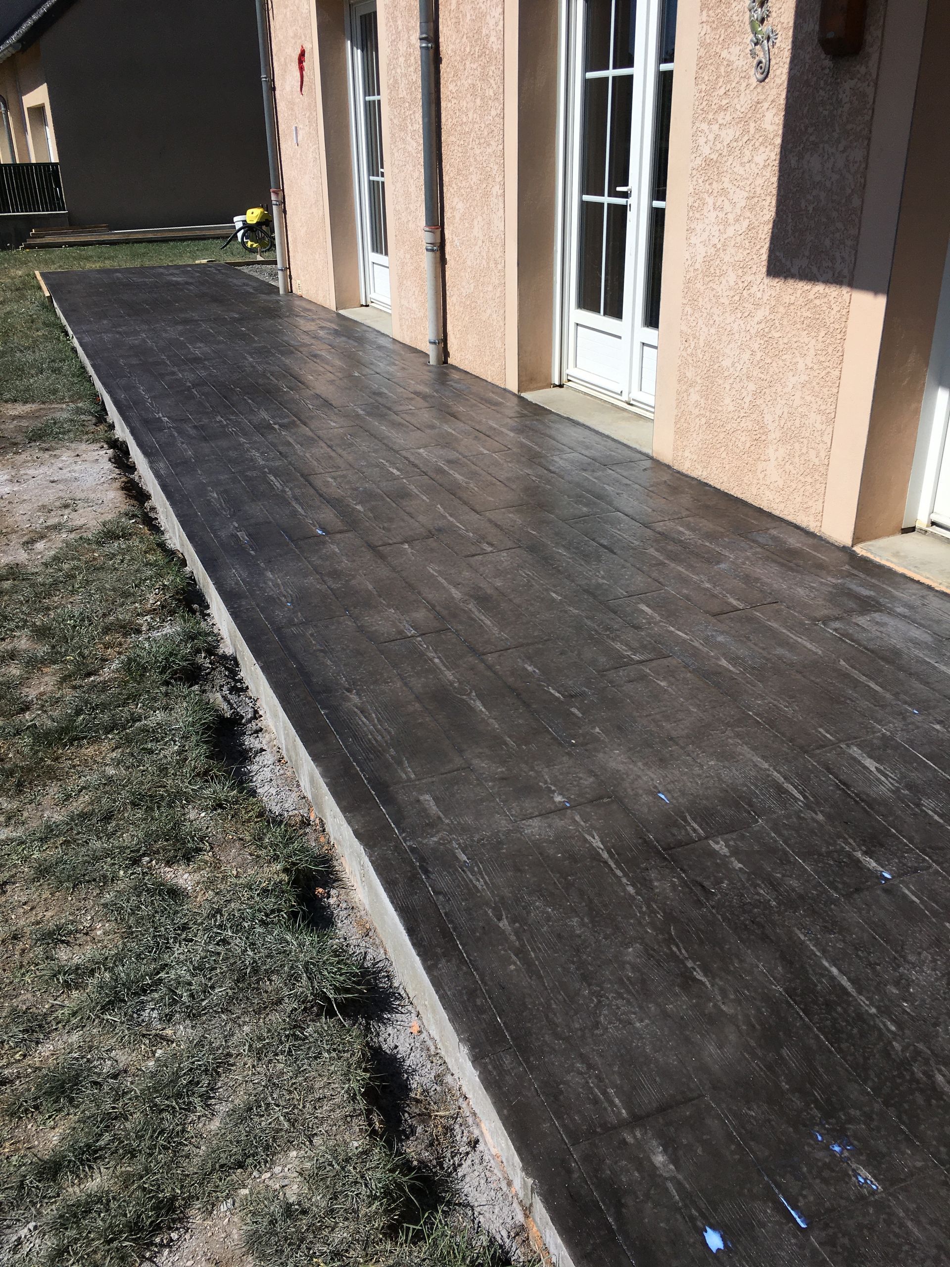 Allée en béton longeant une maison, avec des fenêtres et une porte blanche sur un mur en stuc.