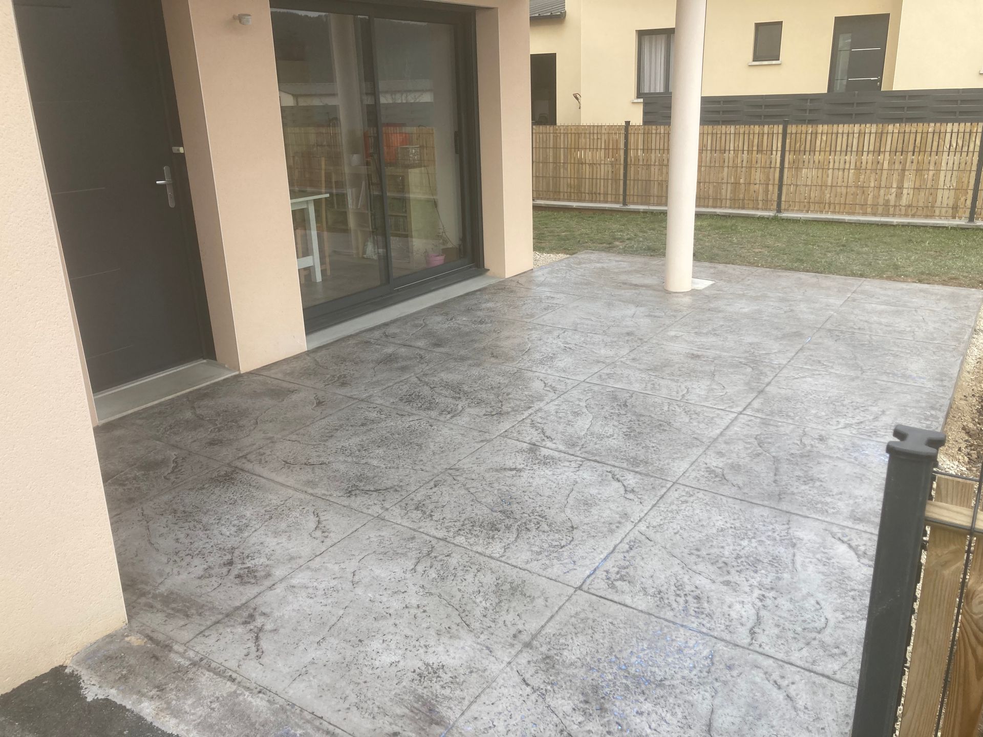 Terrasse couverte avec sol carrelé gris, murs beiges, porte vitrée et balustrade donnant sur un jardin gazonné.