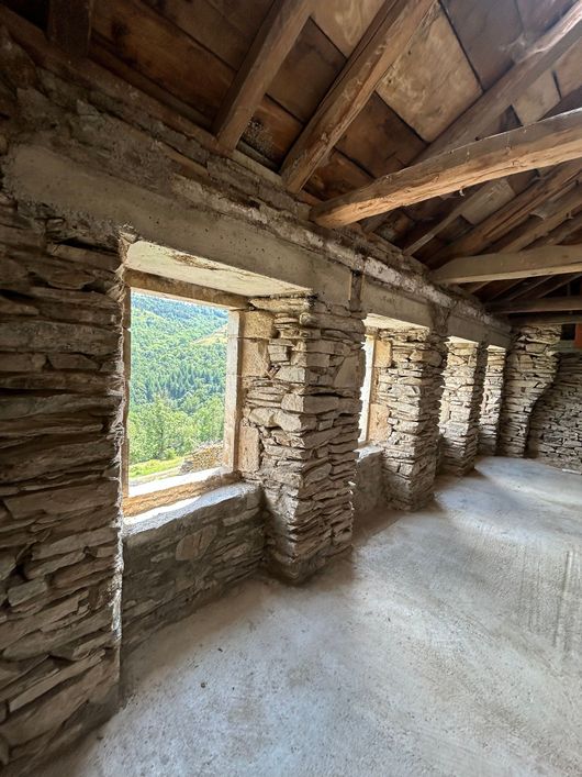 Pièce rustique en pierre avec poutres en bois, une petite fenêtre et une lumière du soleil vive sur le sol.