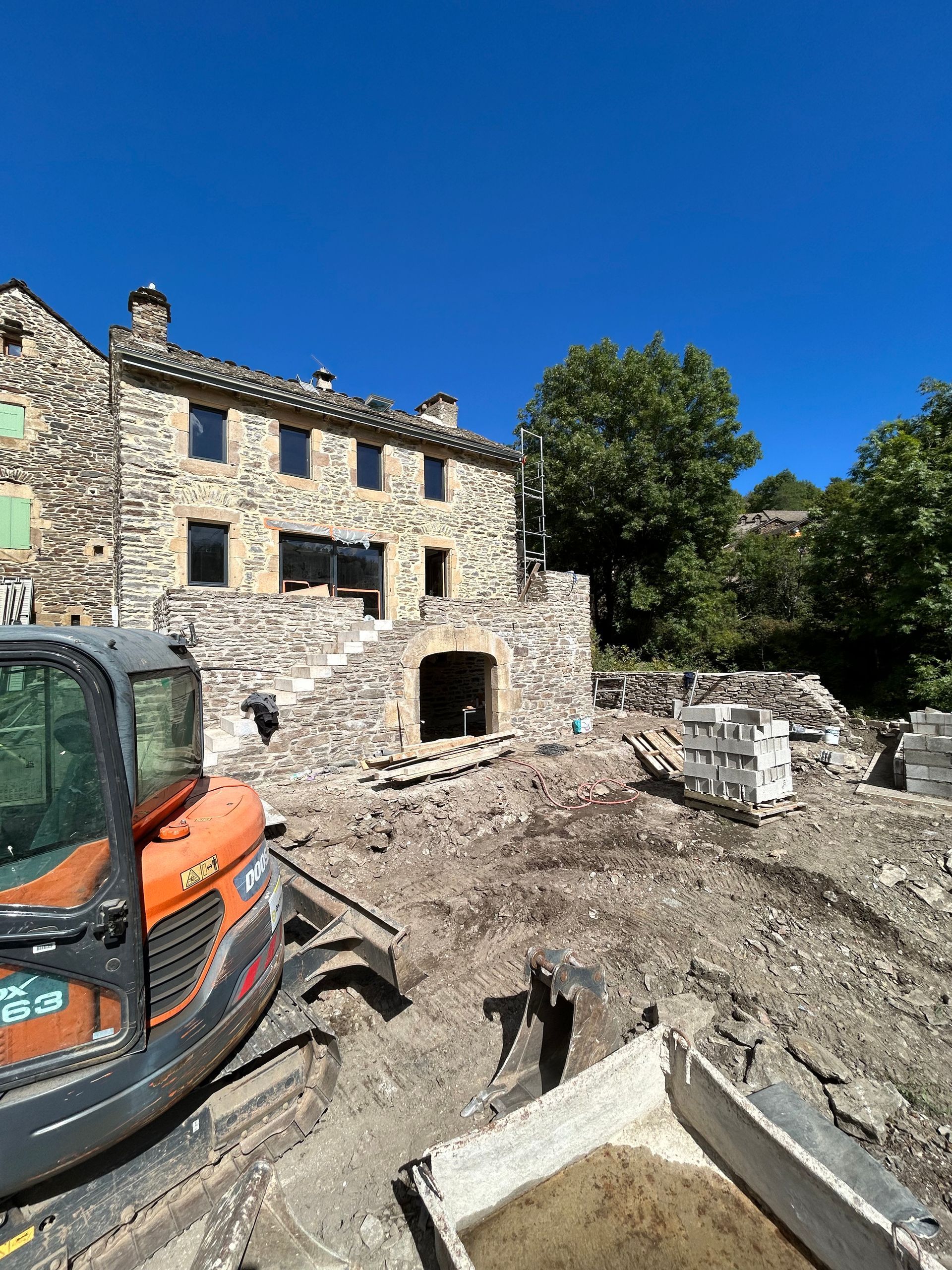 Chantier de démolition avec maison en pierre, pelleteuse, gravats et ciel bleu