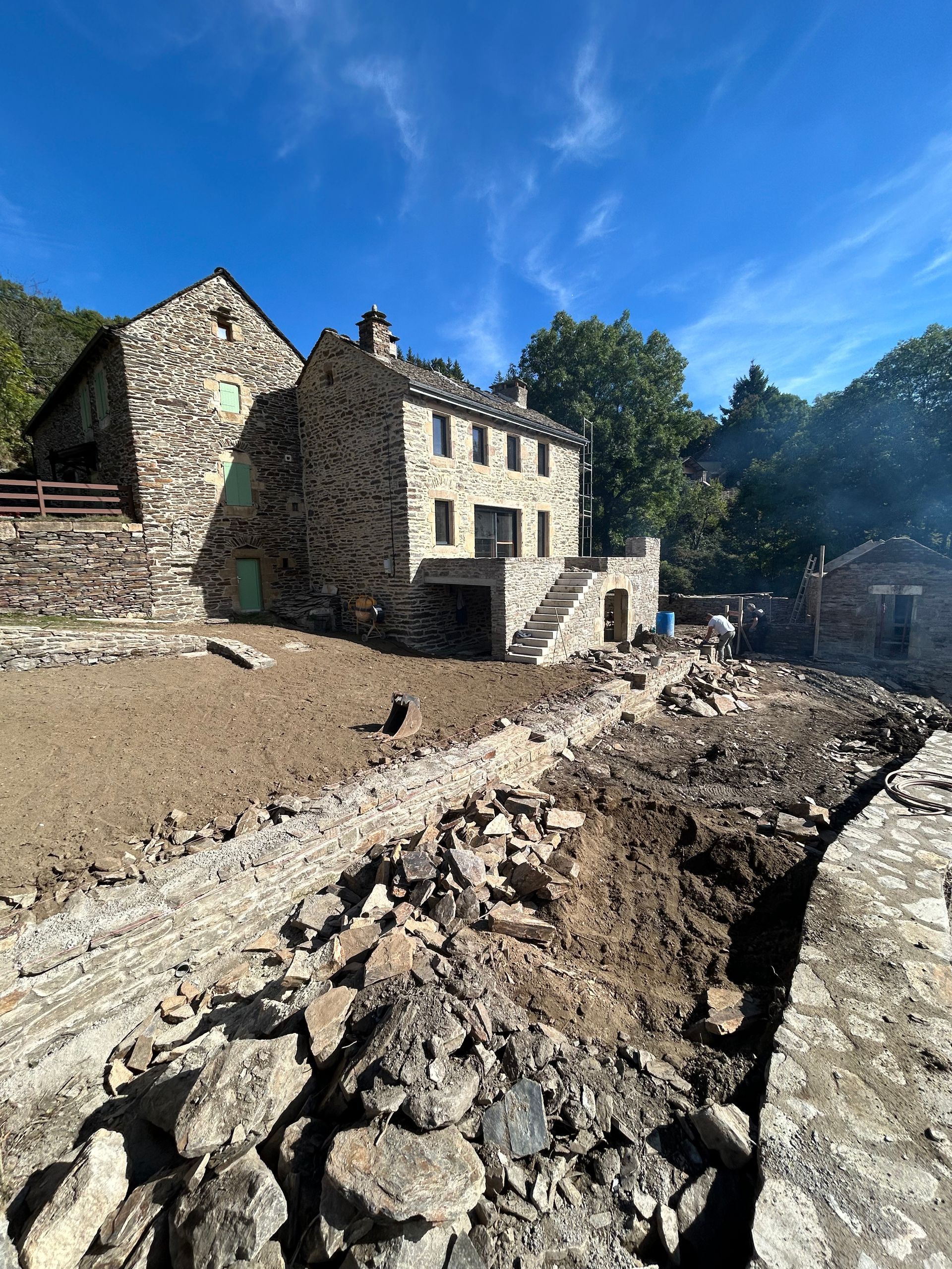 Maison en pierre en rénovation à côté d'une tranchée boueuse, sous un ciel d'un bleu éclatant.