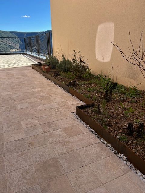 Terrasse ensoleillée avec allée carrelée, mur beige, jardinières et balustrade en verre donnant sur l'eau.
