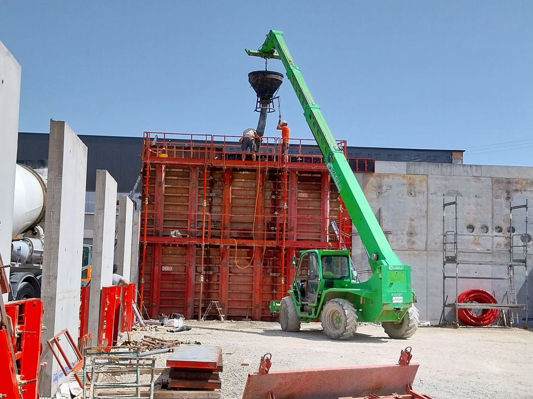 Une grue verte soulève des matériaux à côté d'une structure d'échafaudage rouge sur un chantier de construction.