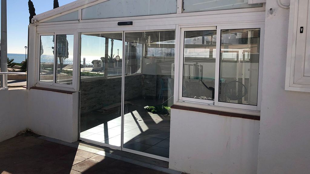 Terraza acristalada con puertas y ventanas corredizas de cristal, marco blanco, mucha luz natural, vista exterior.