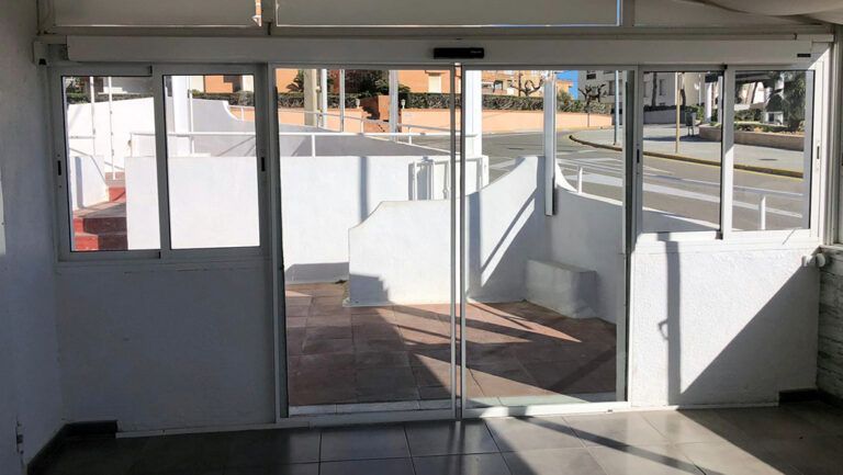 Puertas corredizas automáticas de vidrio, paredes blancas y una rampa conducen a una calle exterior.