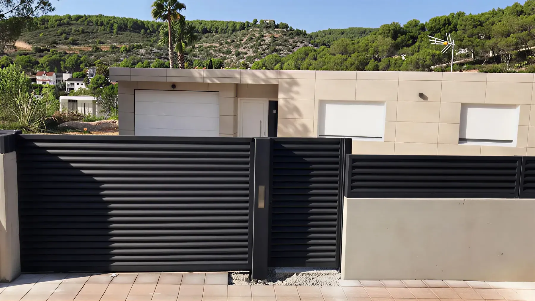 Puerta de entrada con listones negros frente a un edificio moderno de color beige con puerta de garaje y ventanas blancas, contra una ladera.