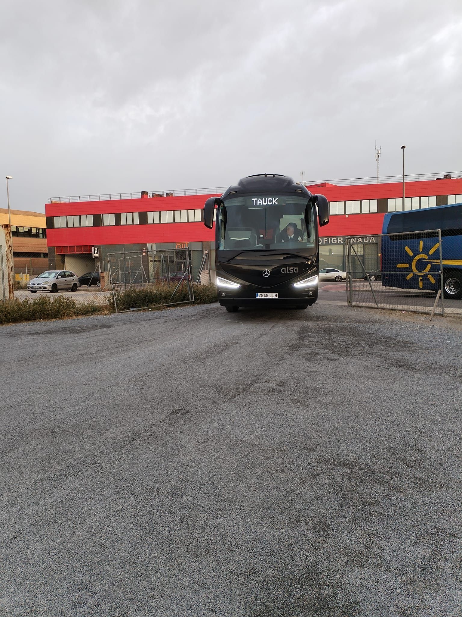 Un autobús negro está estacionado en un estacionamiento de grava frente a un edificio.