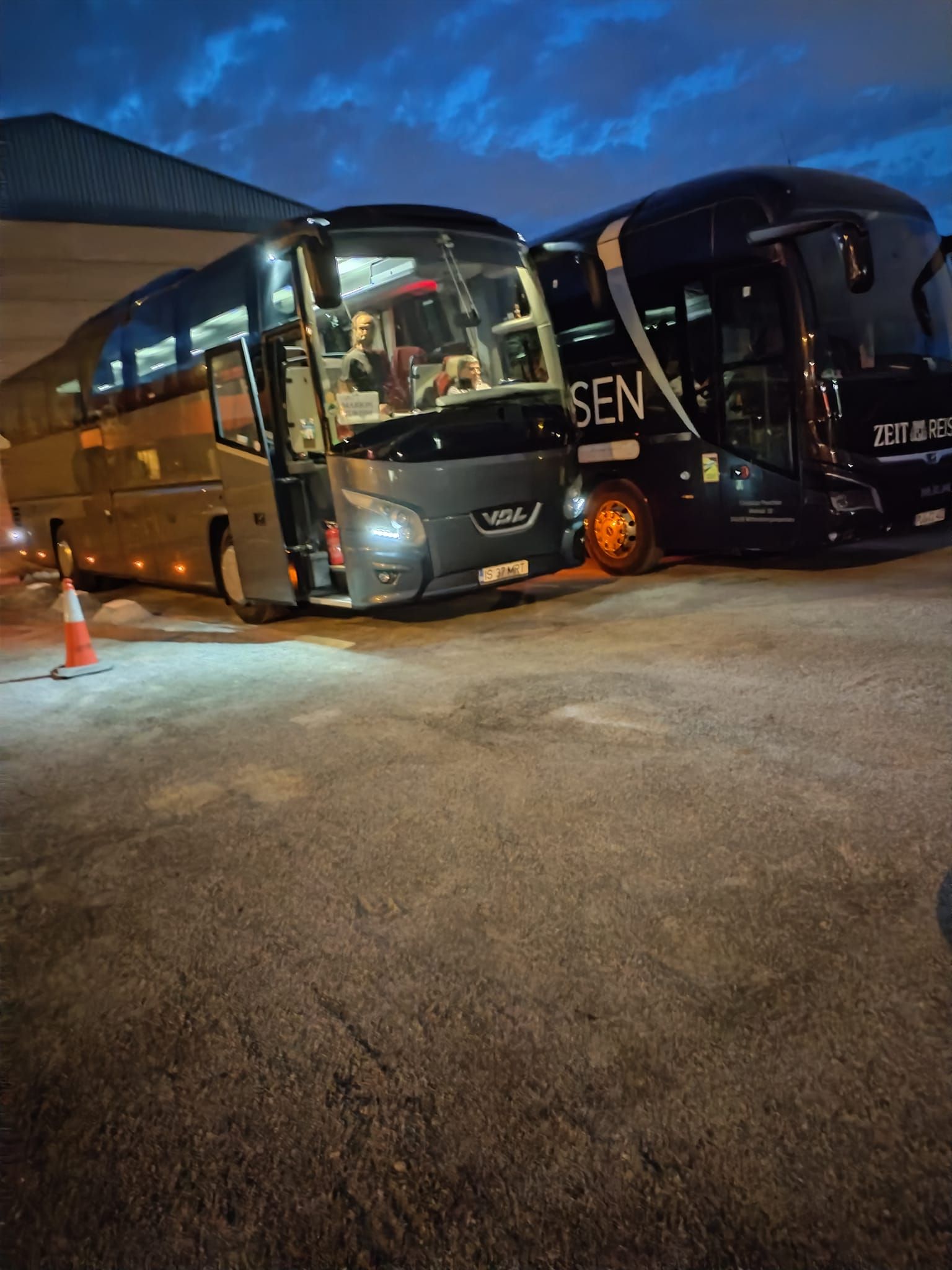 Dos autobuses están estacionados uno al lado del otro en un estacionamiento por la noche.
