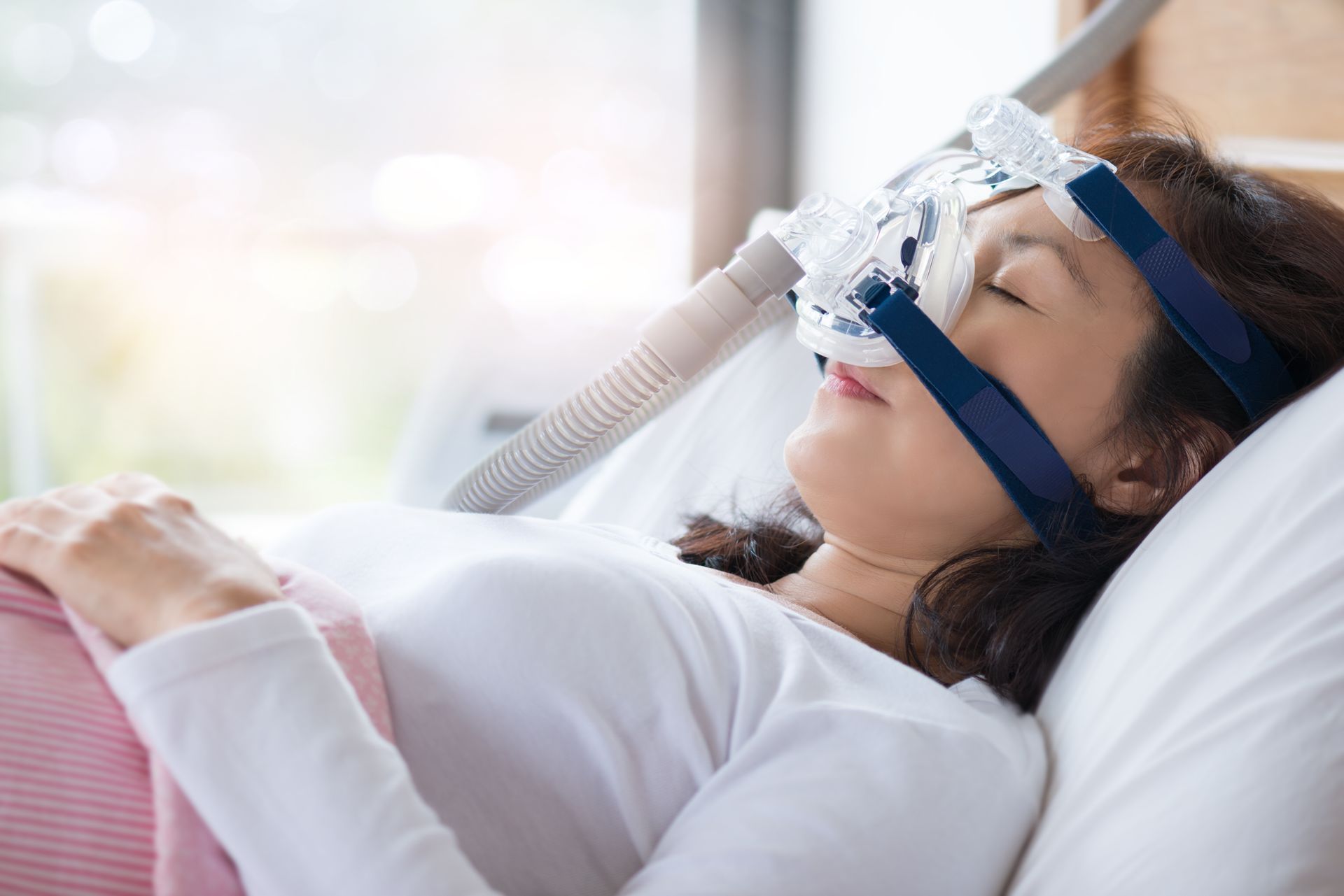 Eine Frau mit CPAP-Maske schläft auf einem Bett in der Nähe eines hellen Fensters.