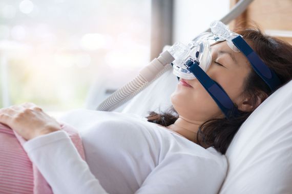 Eine Person mit CPAP-Maske liegt mit geschlossenen Augen im Bett und ruht sich an einem hellen Fenster aus.