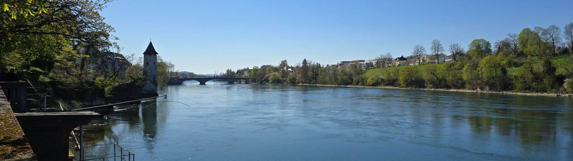 Eine Flusslandschaft mit Turm, Brücke und Gebäuden unter klarem, blauem Himmel.