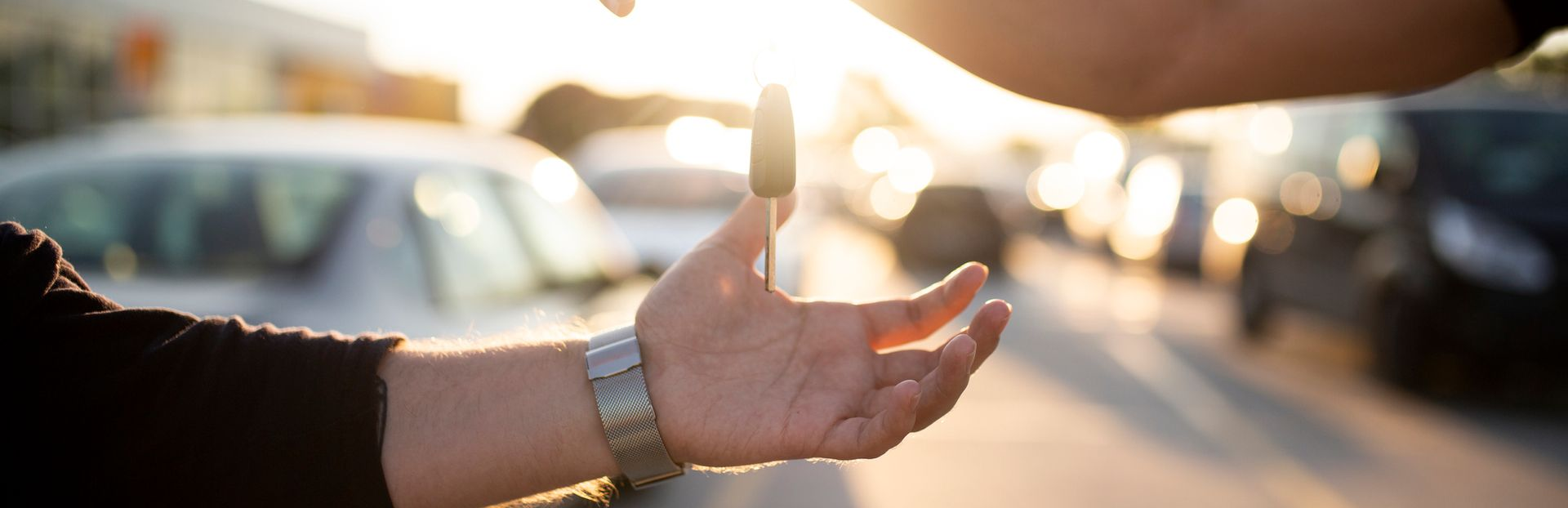 Mano recibiendo llaves de coche de otra mano, con coches borrosos en el fondo, luz solar brillante.