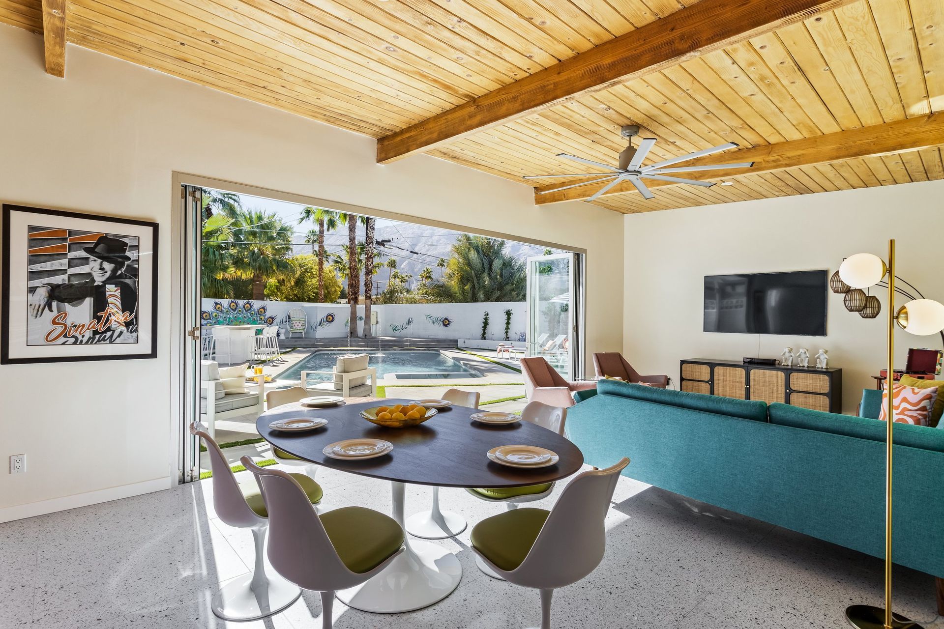 Living room with pool view: round dining table, turquoise sofa, wooden ceiling, open sliding glass doors.