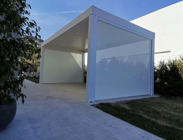 Pérgola blanca con mosquiteras retráctiles, sobre patio de hormigón, junto a zona verde.