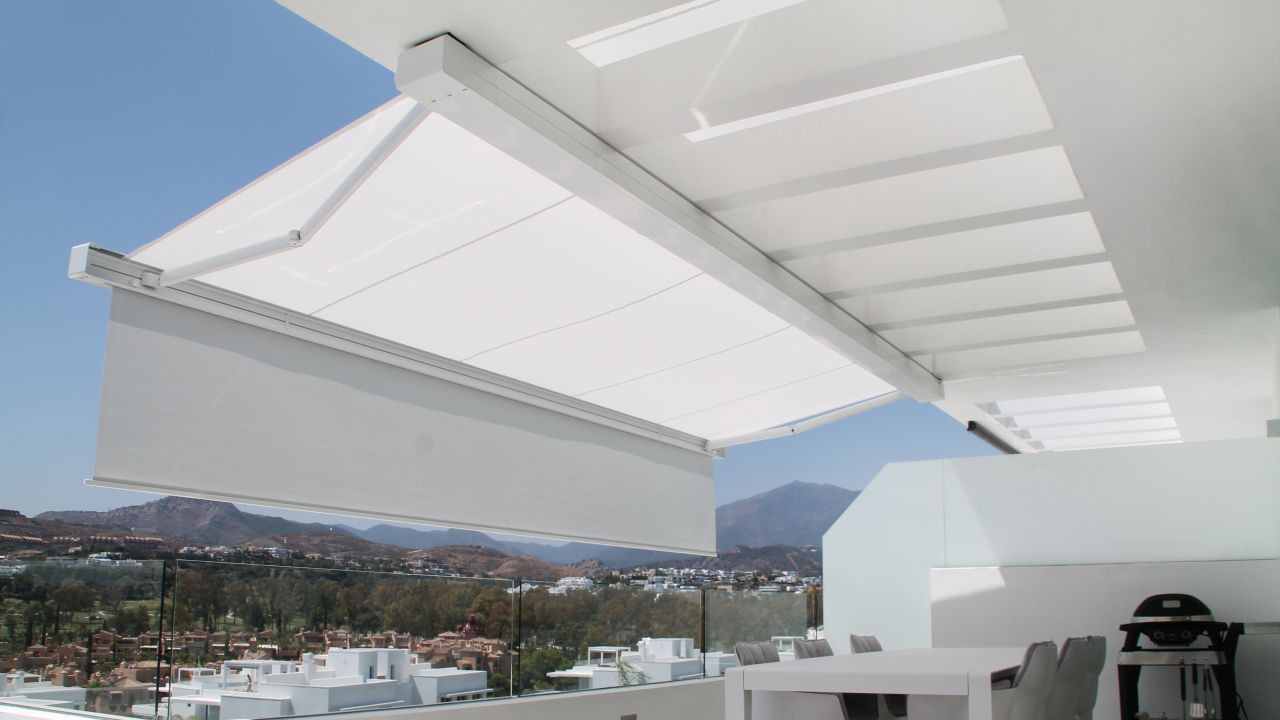 Cafetería al aire libre con mesas y sillas bajo un toldo retráctil; suelo mojado visible.
