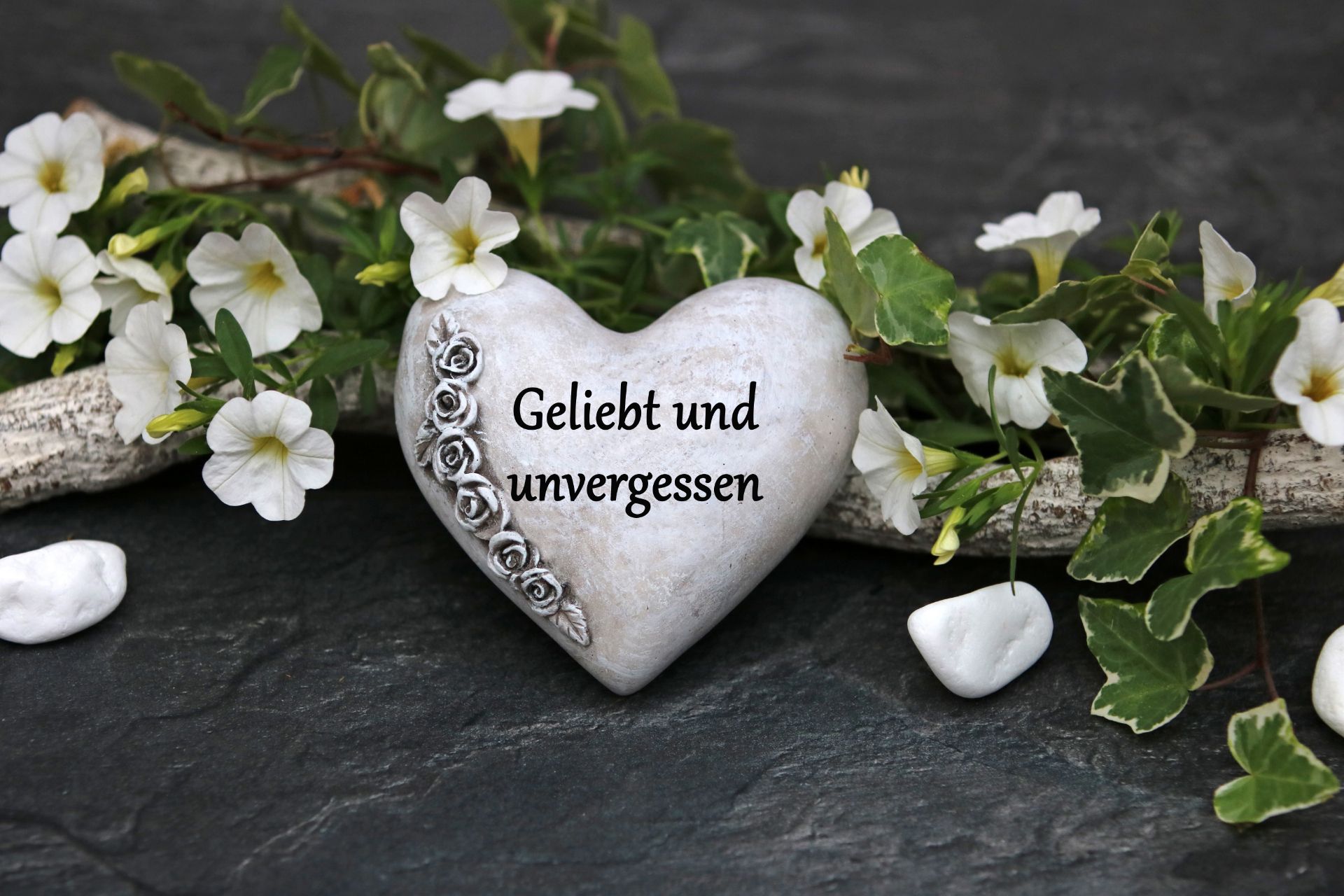 Herzförmiger Stein mit der Aufschrift „Geliebt und unvergessen“ Herzförmiger Stein mit der Aufschrift „Geliebt und unvergessen“, umgeben von weißen Blumen und Grün.