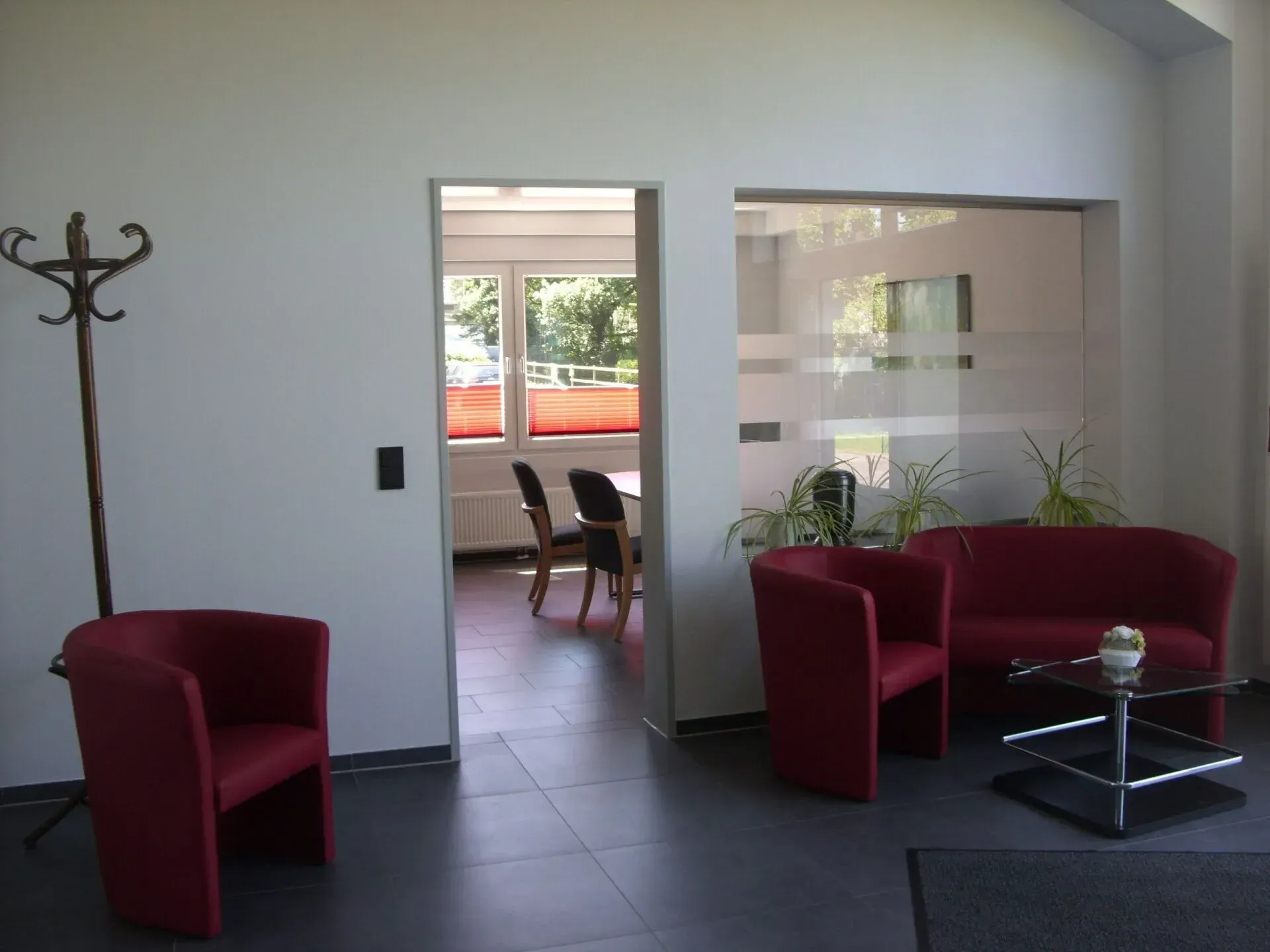 Ein Wartebereich im Büro mit roten Sitzgelegenheiten, einer Garderobe und einem teilweise einsehbaren Konferenzraum.