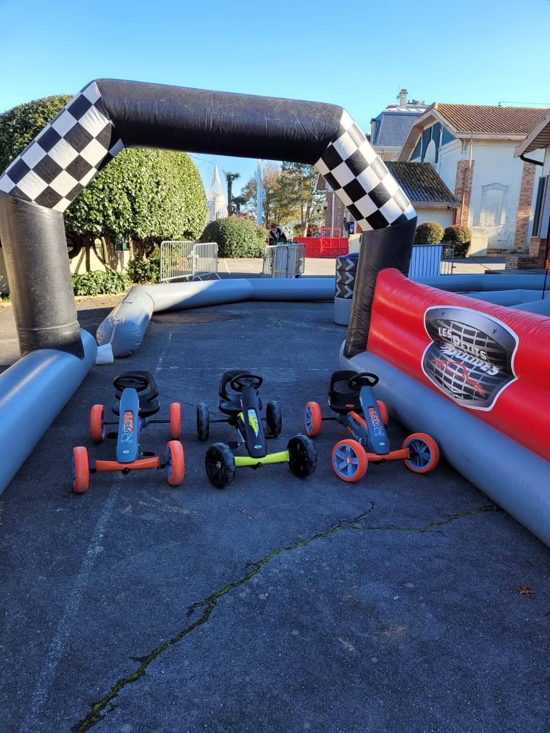 Trois karts à pédales alignés sur une piste de course gonflable pour enfants. Une arche d'arrivée en damier surplombe la piste. Journée ensoleillée.