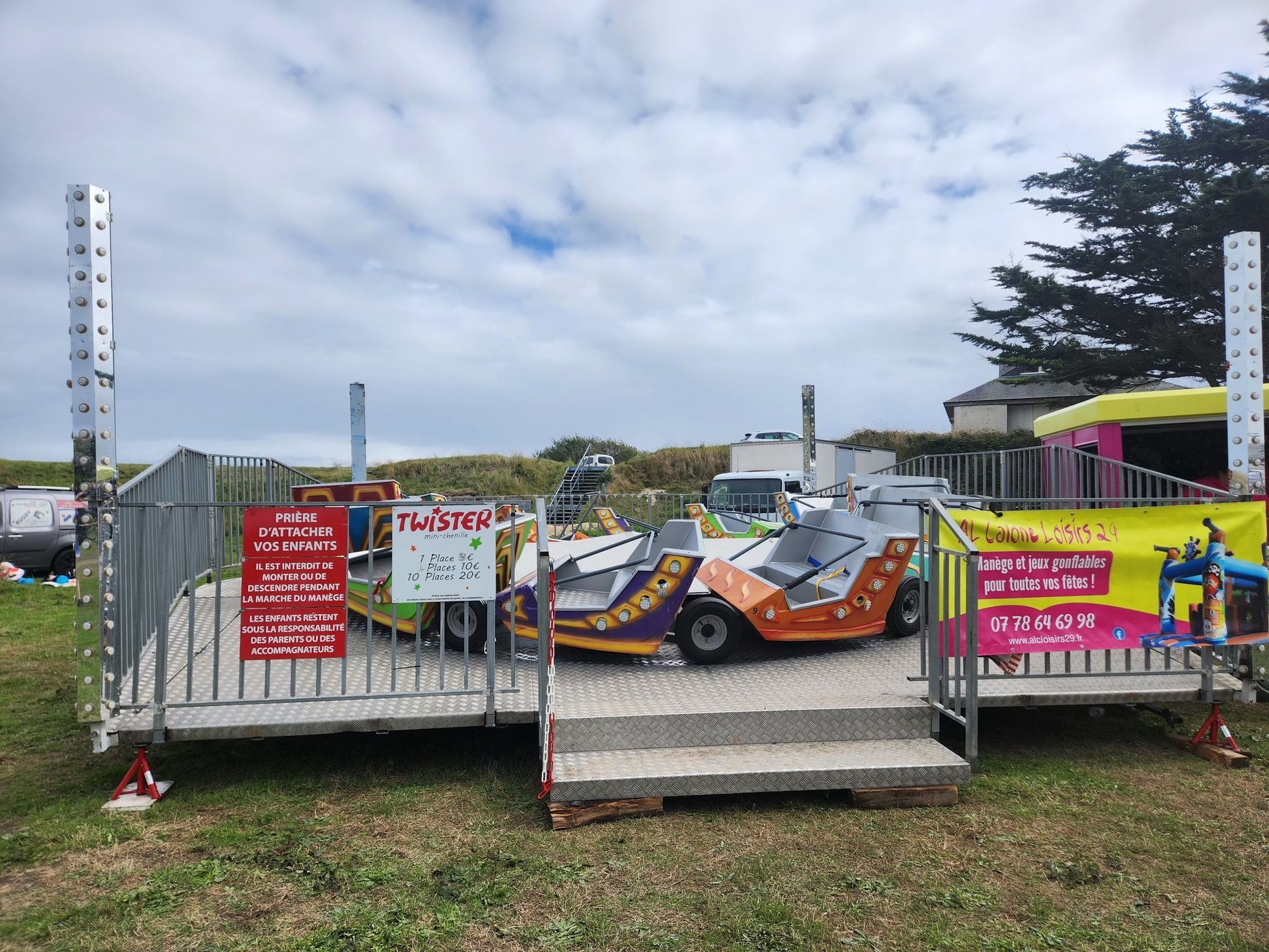 Manège de carnaval : voitures de course colorées sur une plateforme entourée de grillages métalliques, sur un terrain herbeux. Ciel nuageux.