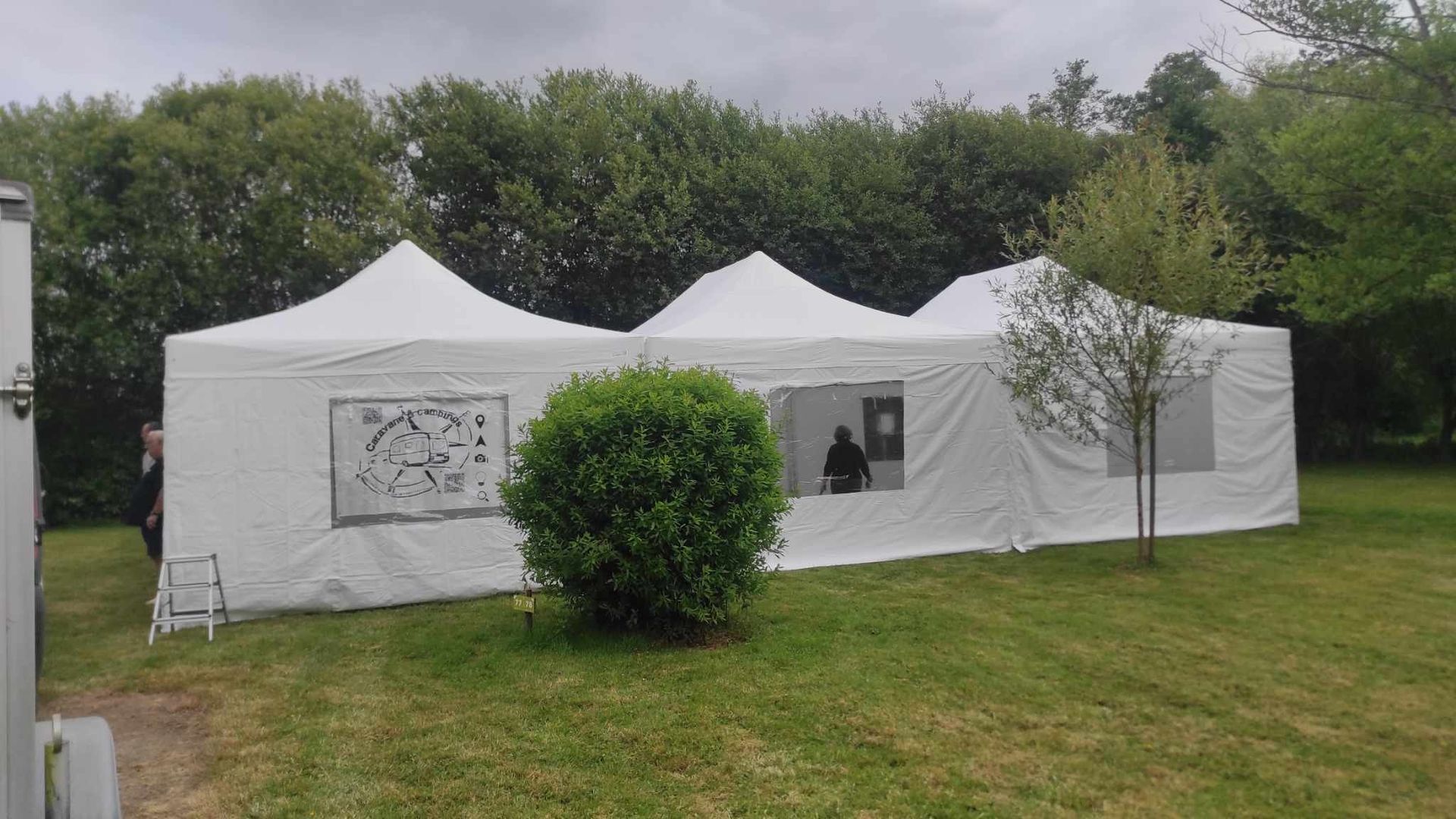 Tente blanche sur l'herbe verte avec des arbres en arrière-plan. Une fenêtre montre une personne à l'intérieur, et un buisson se dresse devant.