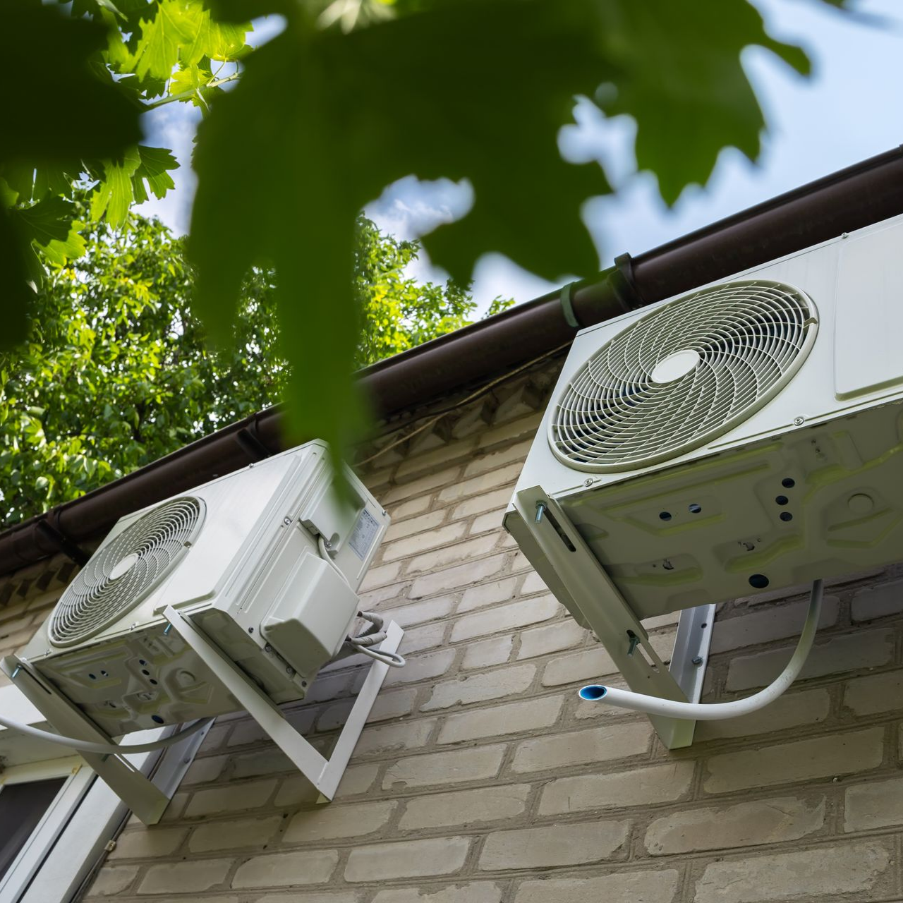 Deux climatiseurs muraux sur un bâtiment en briques, sous des feuilles d'arbre vertes