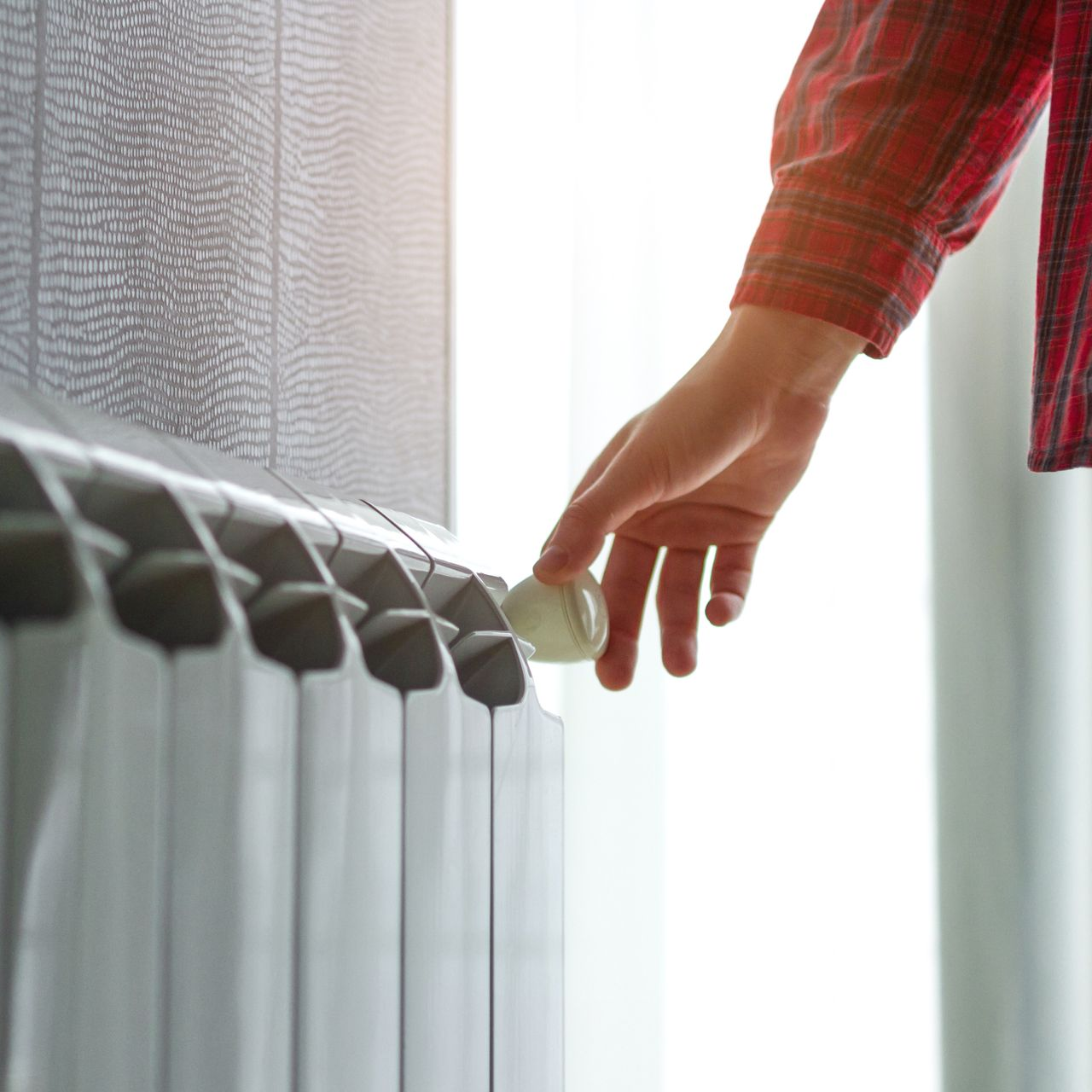 Une main actionne un robinet de radiateur près d'une fenêtre, laissant apparaître une manche à carreaux rouges.
