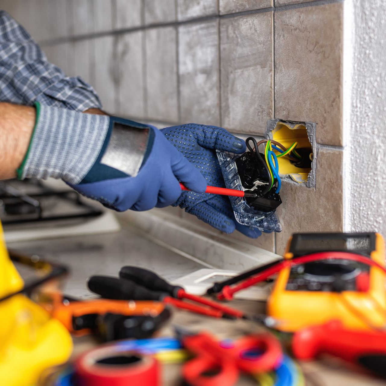 Un électricien installe une prise murale, portant des gants bleus et ses outils sur un établi à proximité.
