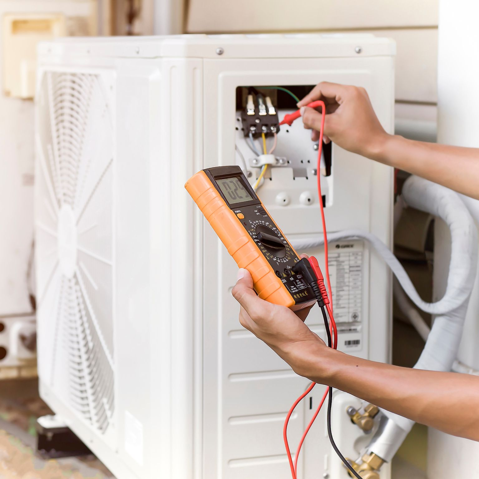 Un technicien teste le câblage d'un système de chauffage, ventilation et climatisation à l'aide d'un multimètre sur une unité extérieure blanche.