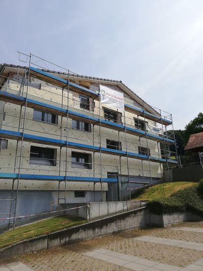 Gebäude im Bau, Gerüste verhüllen die Außenseite. Das Gebäude ist beige mit dunklen Fensterrahmen.