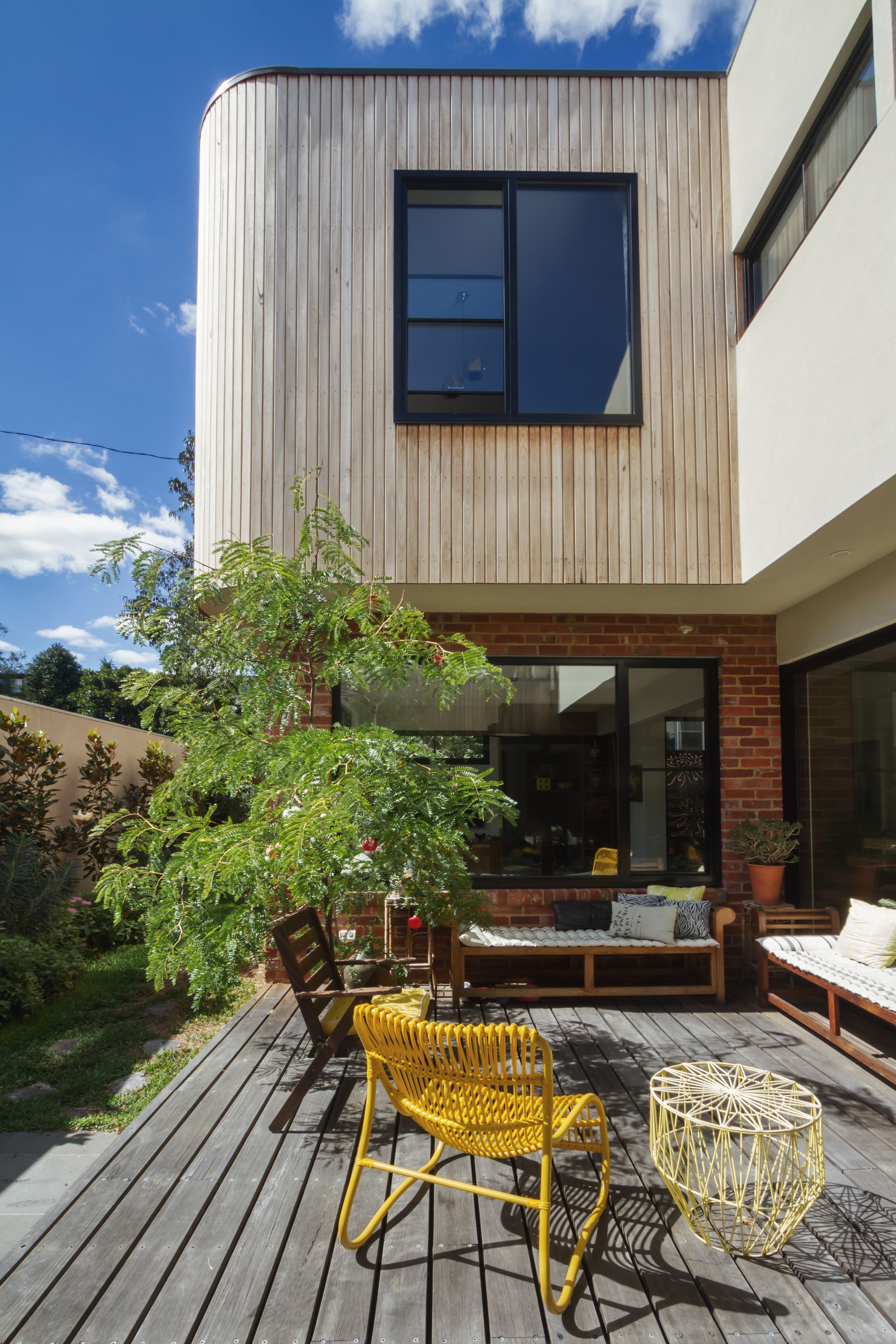 Un patio avec une chaise jaune, un banc en bois et un petit arbre, adossé à une maison moderne.