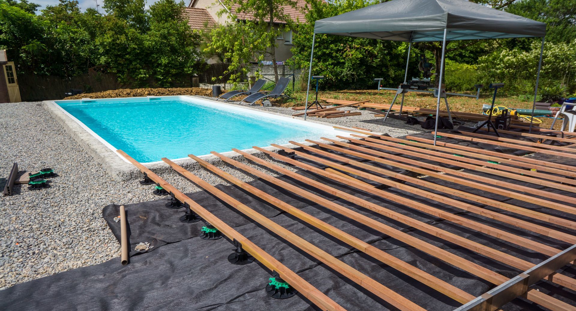 Une piscine dans un jardin, avec des lames de terrasse en bois posées sur une structure recouverte d'une bâche de protection.