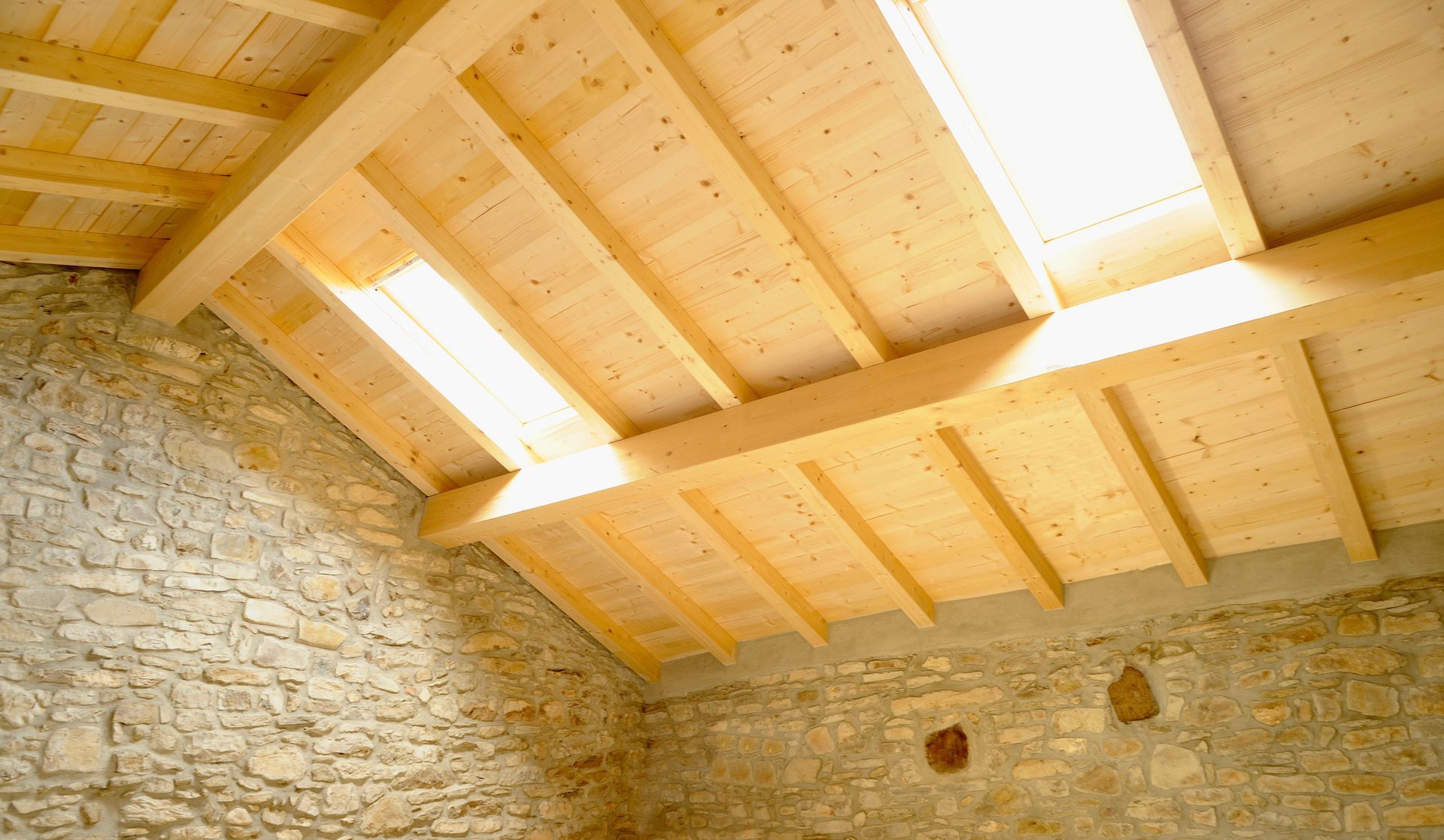 Un plafond en bois incliné avec des poutres apparentes et deux puits de lumière au-dessus d'un mur en pierre.
