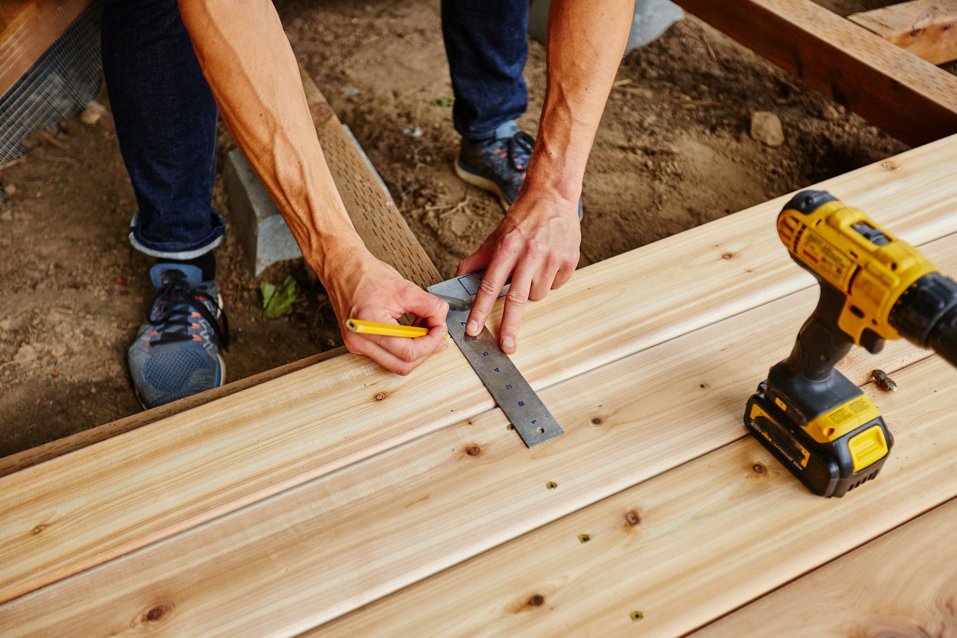 Une personne utilise une équerre et un crayon pour tracer une ligne sur des planches de terrasse en bois.