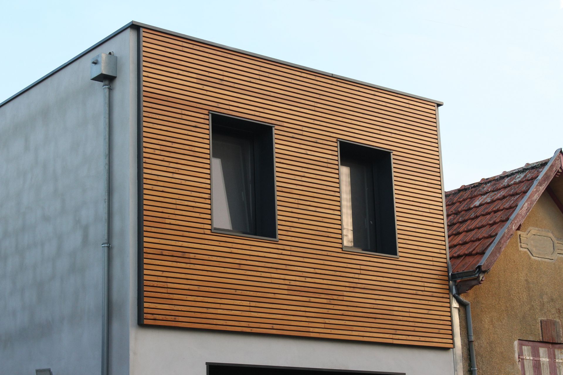 La façade d'un bâtiment de deux étages présente un revêtement en lattes de bois horizontales à l'étage et deux fenêtres.